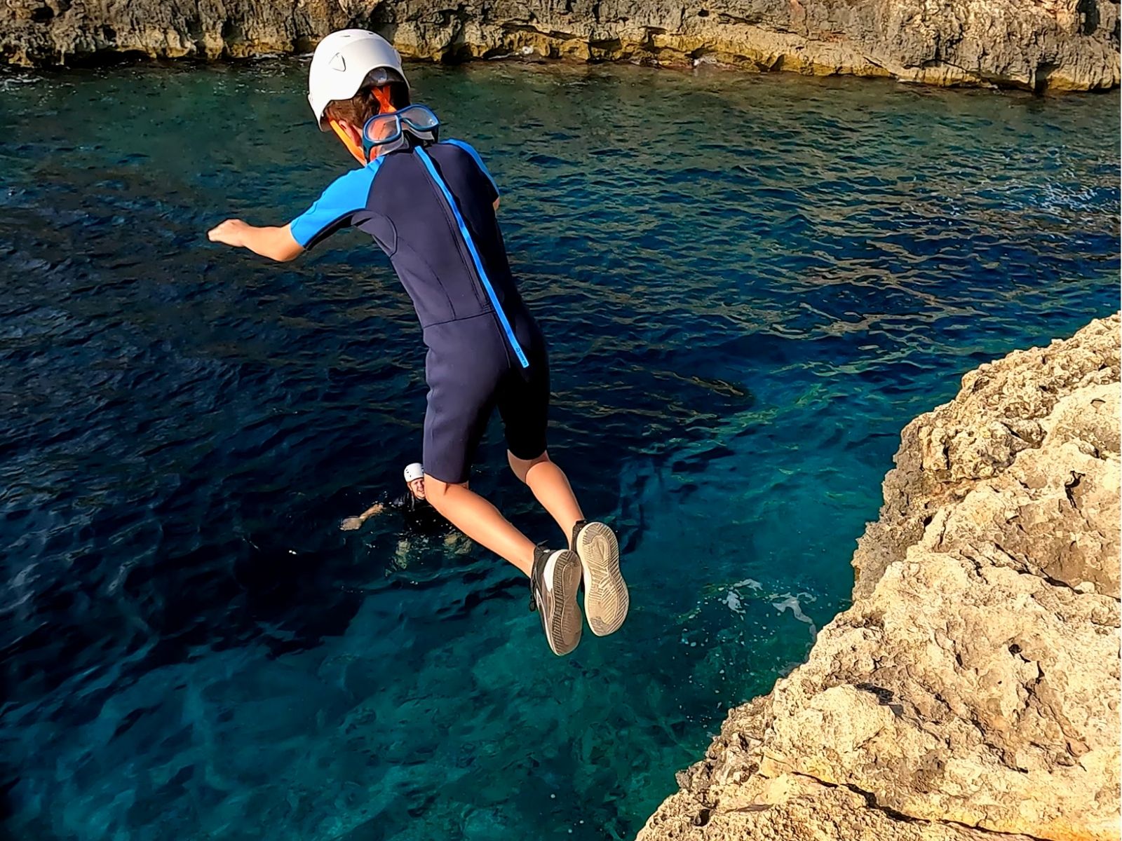 Niño con casco saltando al mar durante una actividad de coasteering, con un guía en el agua